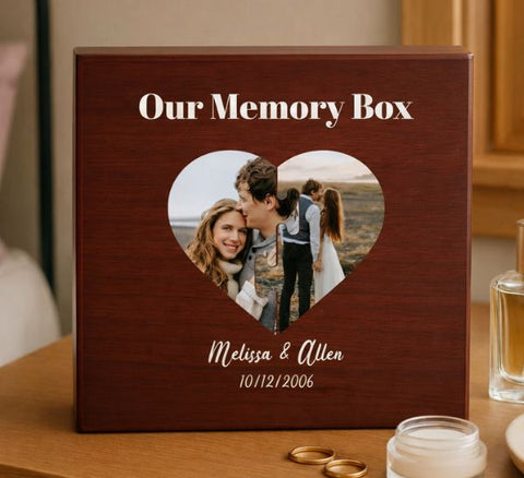 Dark wood memory box with heart-shaped photo cutout displaying couple, engraved with names and date, displayed on table with rings and candle.