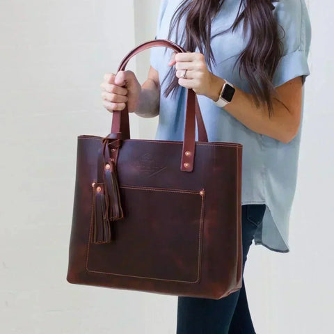 Woman in a light blue shirt holding a dark burgundy leather tote bag with a leather tassel, a front pocket and copper rivets.