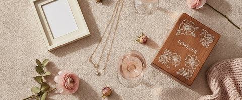 A blush flat-lay of personalized anniversary gifts for her including two gold necklaces, a photo frame, an engraved keepsake box, crystal wine glasses with rose wine, dried roses, and a soft throw.