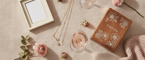 A blush flat-lay of personalized anniversary gifts for her with two gold necklaces, a photo frame, an engraved keepsake box, crystal wine glasses, dried roses and a soft throw.