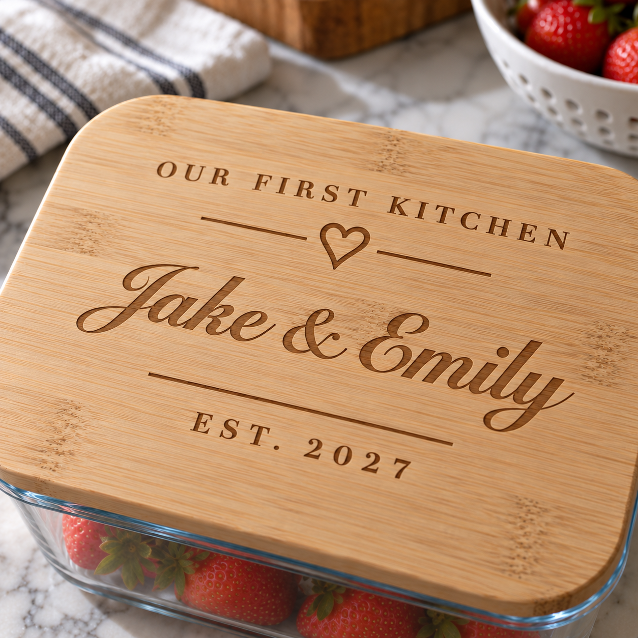 Close-up of a bamboo storage lid engraved Our First Kitchen with a heart, Jake & Emily in script and EST. 2027, on a glass container of strawberries
