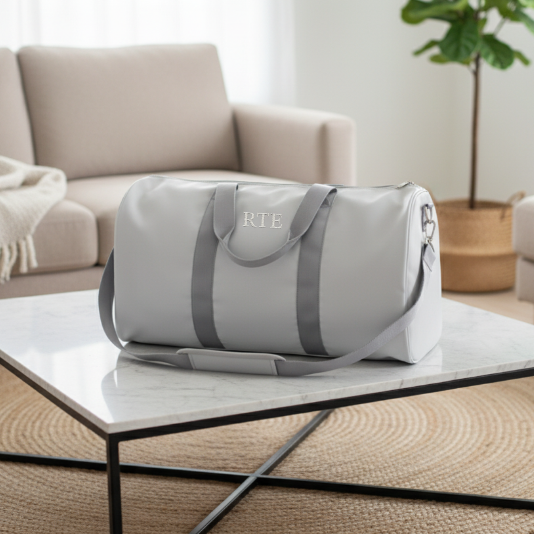 Light grey duffel bag with grey handles and straps, embroidered with sample monogram "RTE" on front panel, displayed on marble table in home setting