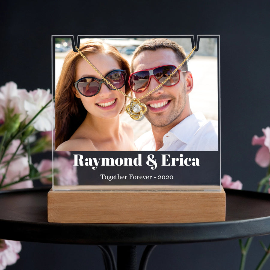 Personalized acrylic frame with wooden base displaying couple photo with sample names "Raymond & Erica" and "Together Forever - 2020" text, paired with gold love knot necklace