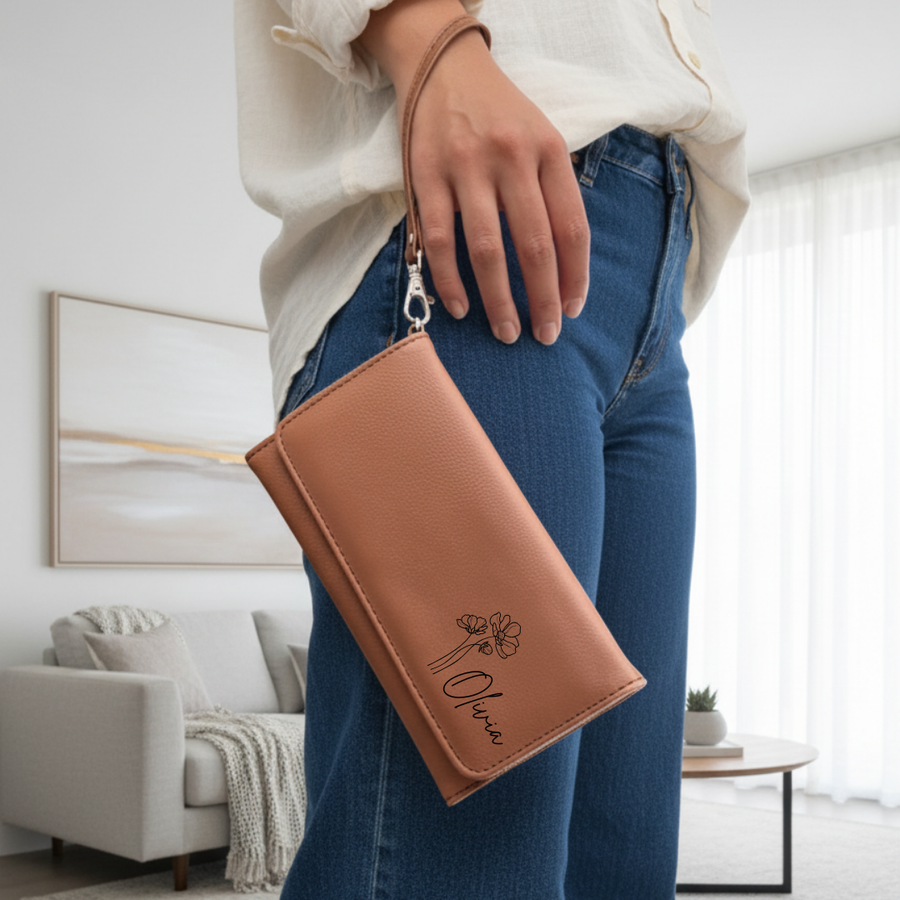 Tan leatherette wristlet with sample birth flower design and name "Olivia" engraved on front, held by woman wearing cream sweater and blue jeans