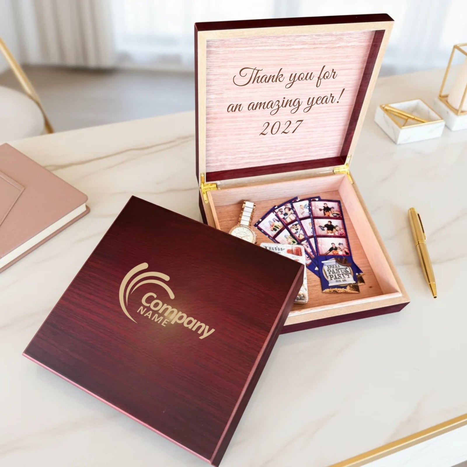 Dark cherry wood keepsake box engraved with sample company logo on lid, open to reveal pink cedar interior with engraved sample message "Thank you for an amazing year! 2027" and contents including watch, photo cards, and medal.