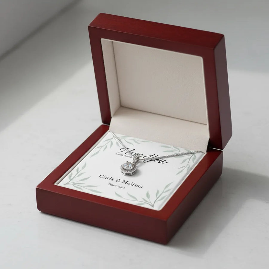 Burgundy gift box with cream interior displaying a silver necklace with cushion-cut pendant, resting on a card personalized with sample names "Chris & Melissa" and "Since 2014