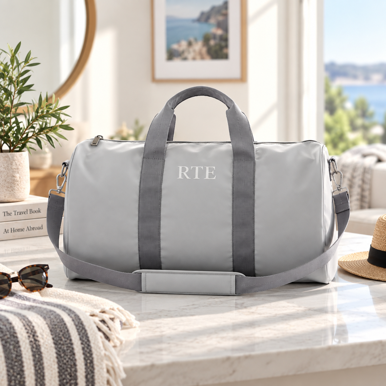 Light grey personalized duffle bag with 'RTE' embroidered in white thread on the front, shown with sunglasses and a straw hat on a marble counter.