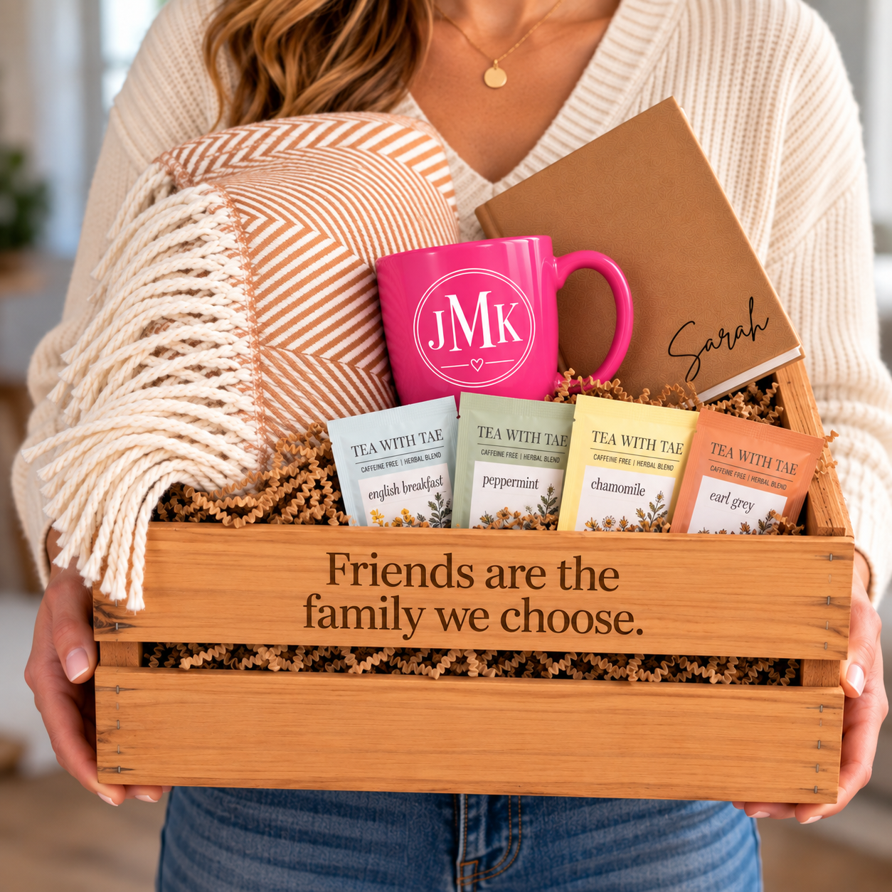 Woman holding a wooden gift crate personalized with a message, filled with a pink monogrammed mug, throw blanket, journal, and tea sachets