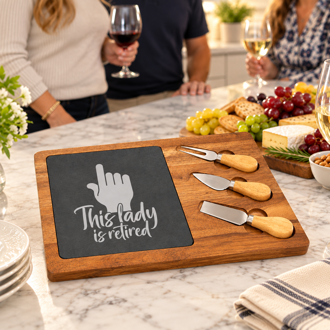 Retirement gift serving board with a laser etched This Lady Is Retired design set on a kitchen counter during a gathering with wine and cheese.