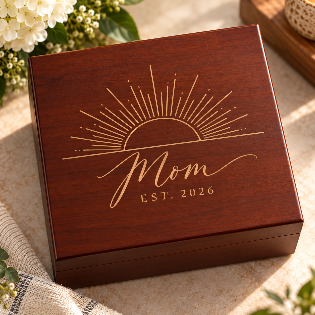 A rectangular wooden keepsake box with a warm reddish-brown finish, featuring an engraved gold sunburst and the words 'Mom EST. 2026' on the lid.