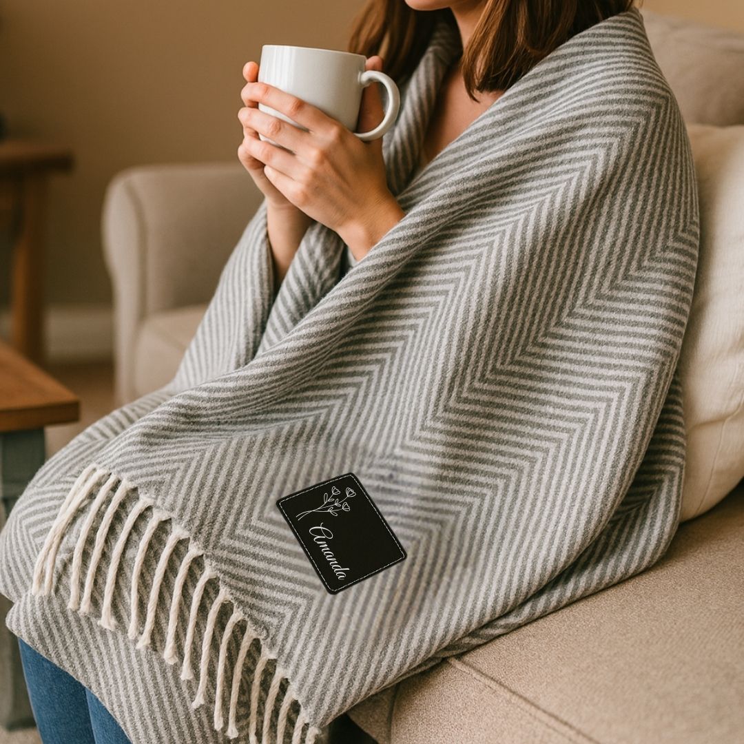 Woman holding white mug while wrapped in gray herringbone throw blanket with fringe trim and black patch embroidered with sample name "Amelia