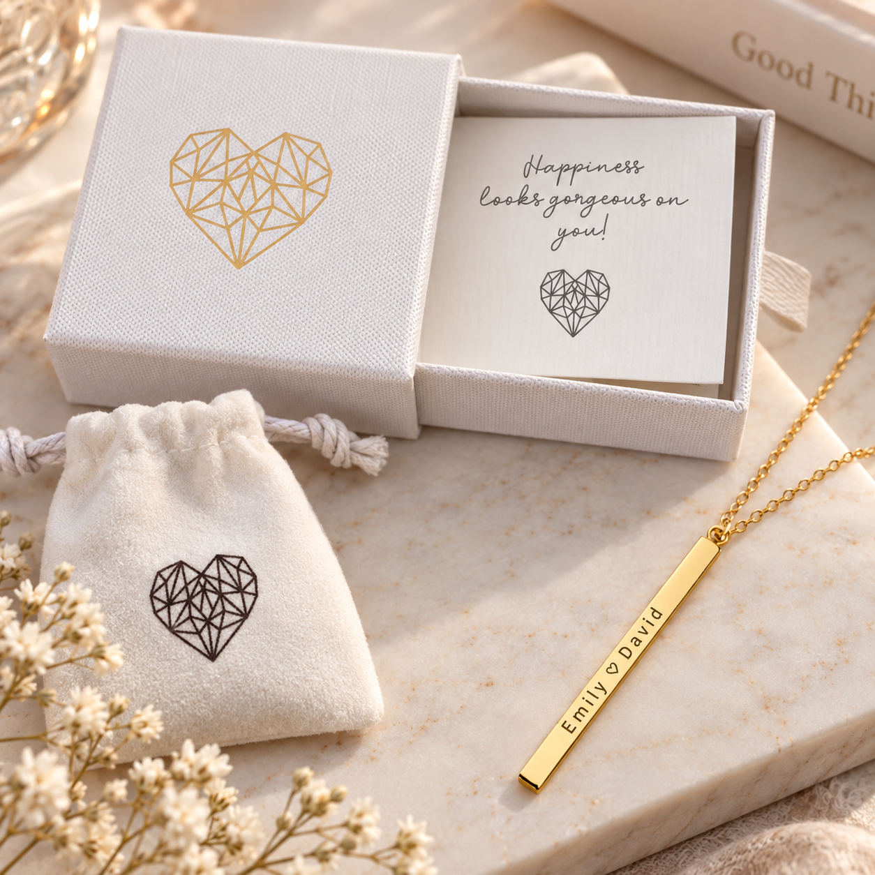 Gold rectangular bar necklace engraved with sample coordinates "Emily 5.30.19", shown with white gift box, pouch, and insert card featuring geometric heart design
