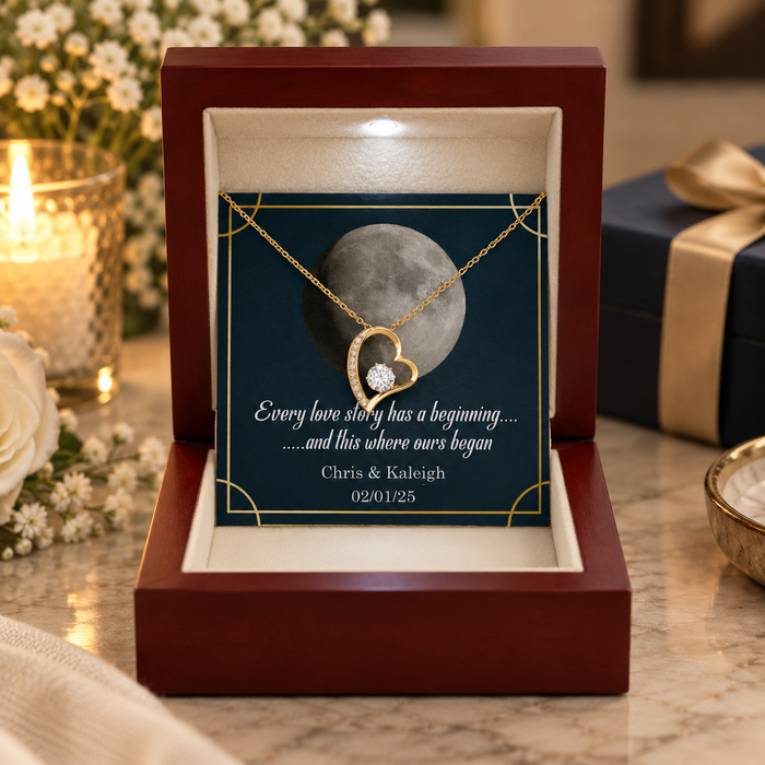 A personalized gold heart pendant necklace displayed inside an open dark red wooden gift box. The necklace is shown over a printed card featuring a moon graphic and custom text, with a lit candle and gift box in the softly blurred background.