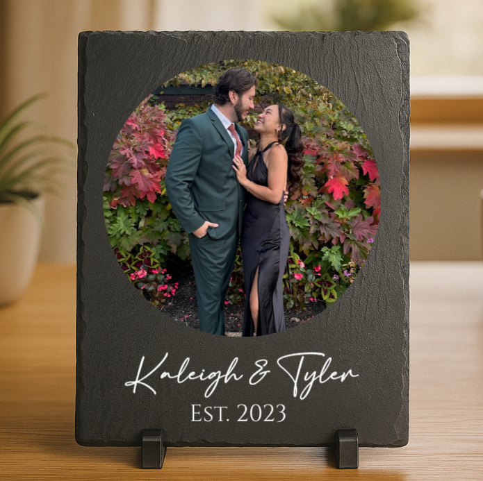 Black slate frame with circular photo cutout showing couple in formal wear before flower arch, engraved with sample names "Kaleigh & Tyler" and "EST. 2023" below