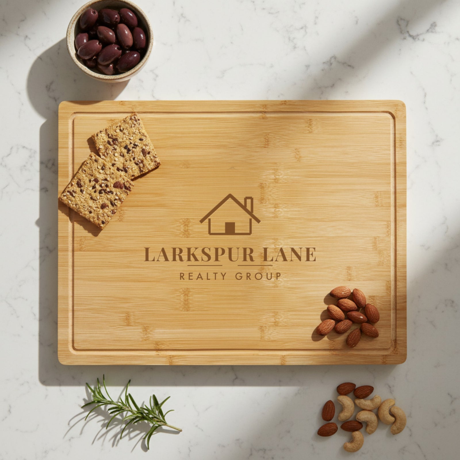 Custom logo cutting board laser engraved with a sample real estate logo, a closing gift for clients shown on a marble counter with olives, crackers and almonds