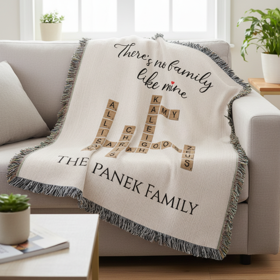 Cream woven blanket draped over a gray sofa, displaying sample family names (Allis, Sarah, Kai, Kamy, Che, Leigh, Zoe, Gus) arranged in crossword-style pattern with "There's no family like mine" text and "THE PANEK FAMILY" at bottom, finished with gray fringe trim.