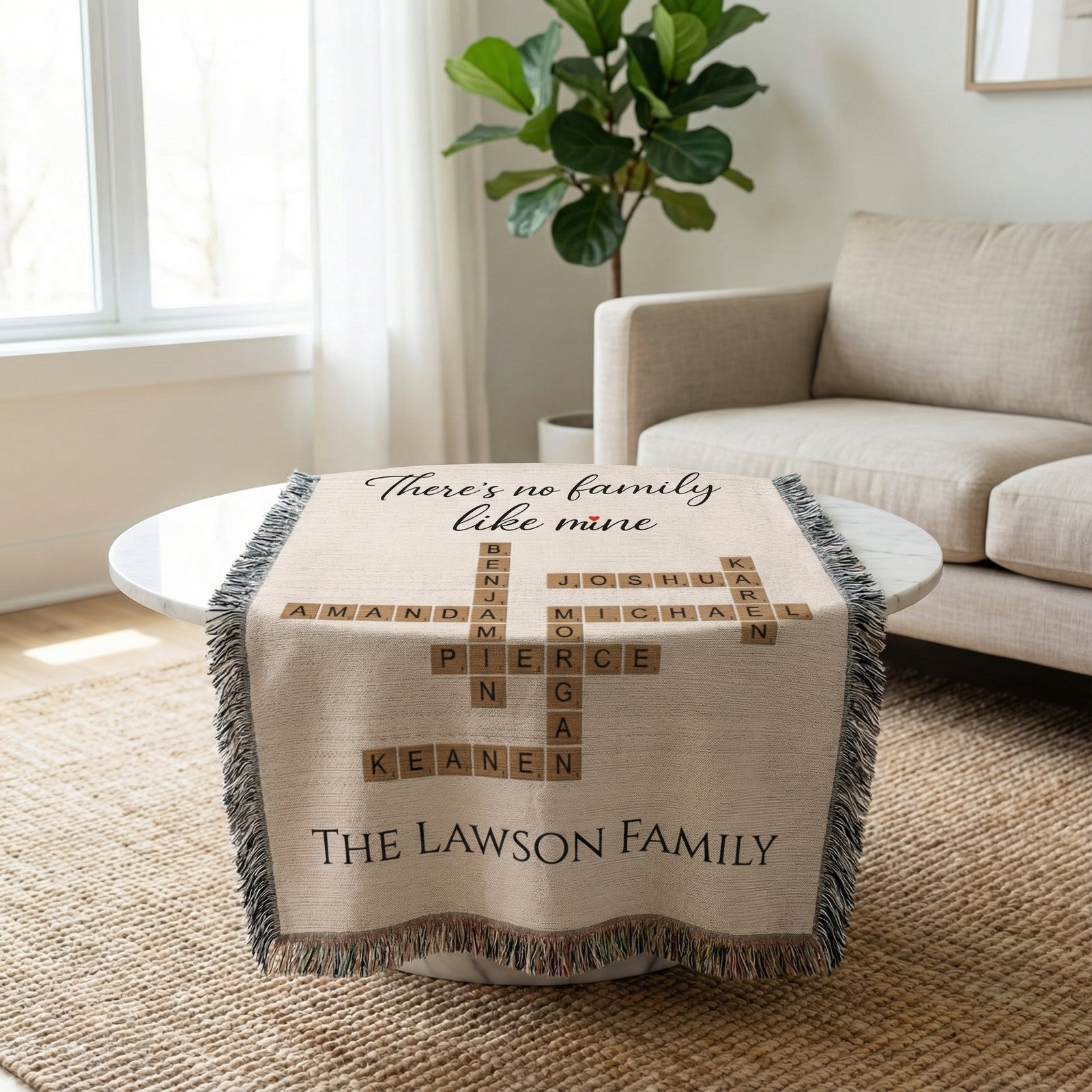 Cream woven blanket with "There's no family like mine" text at top, family names in crossword pattern including samples "Amanda," "Benjamin," "Joshua," "Michael," "Pierce," "Keanen," and "The Lawson Family" at bottom, gray fringe edges, draped over white table in living room.