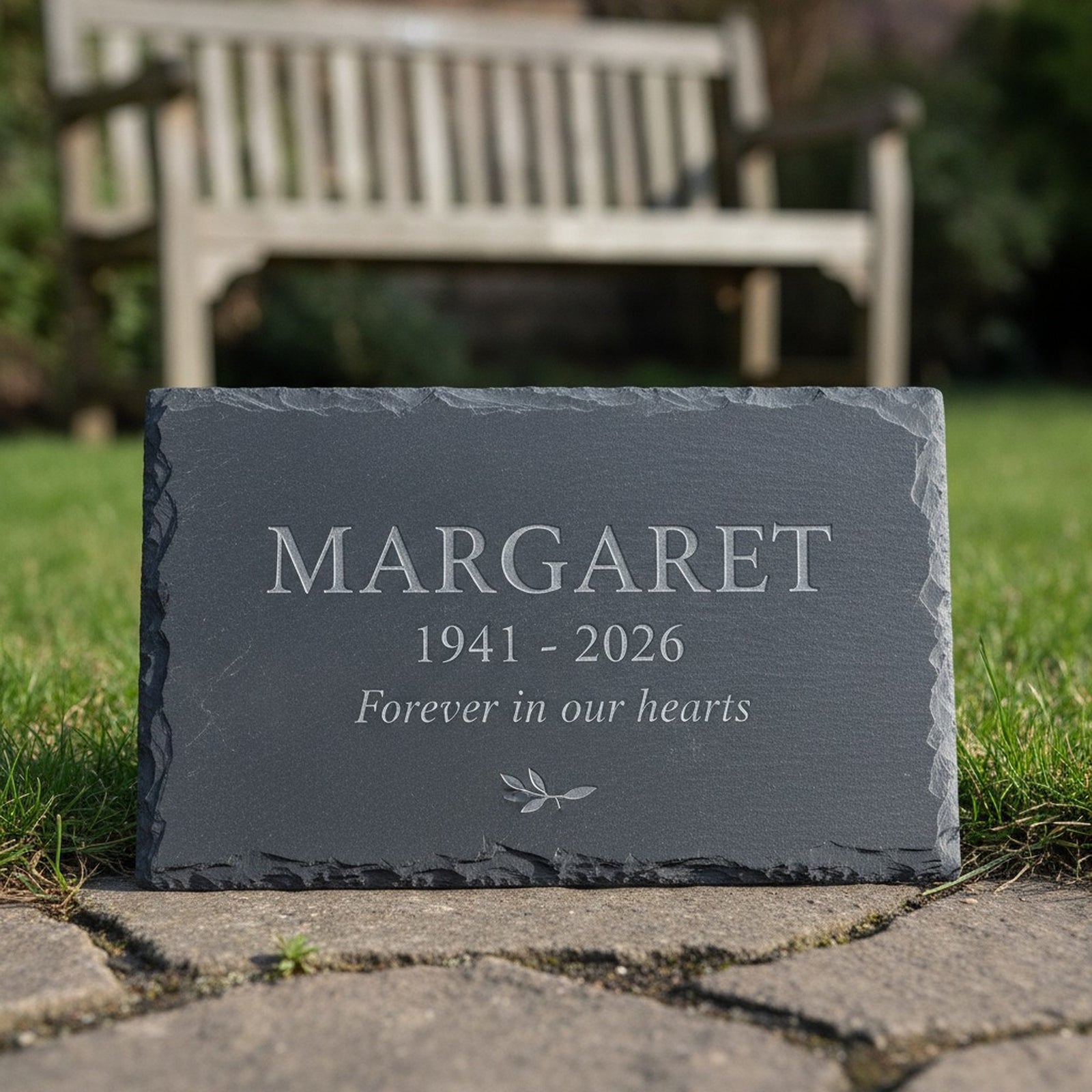 Engraved slate memorial stone resting on grass beside a garden path with a wooden bench behind it, showing the sample name Margaret, the years and a short line beneath