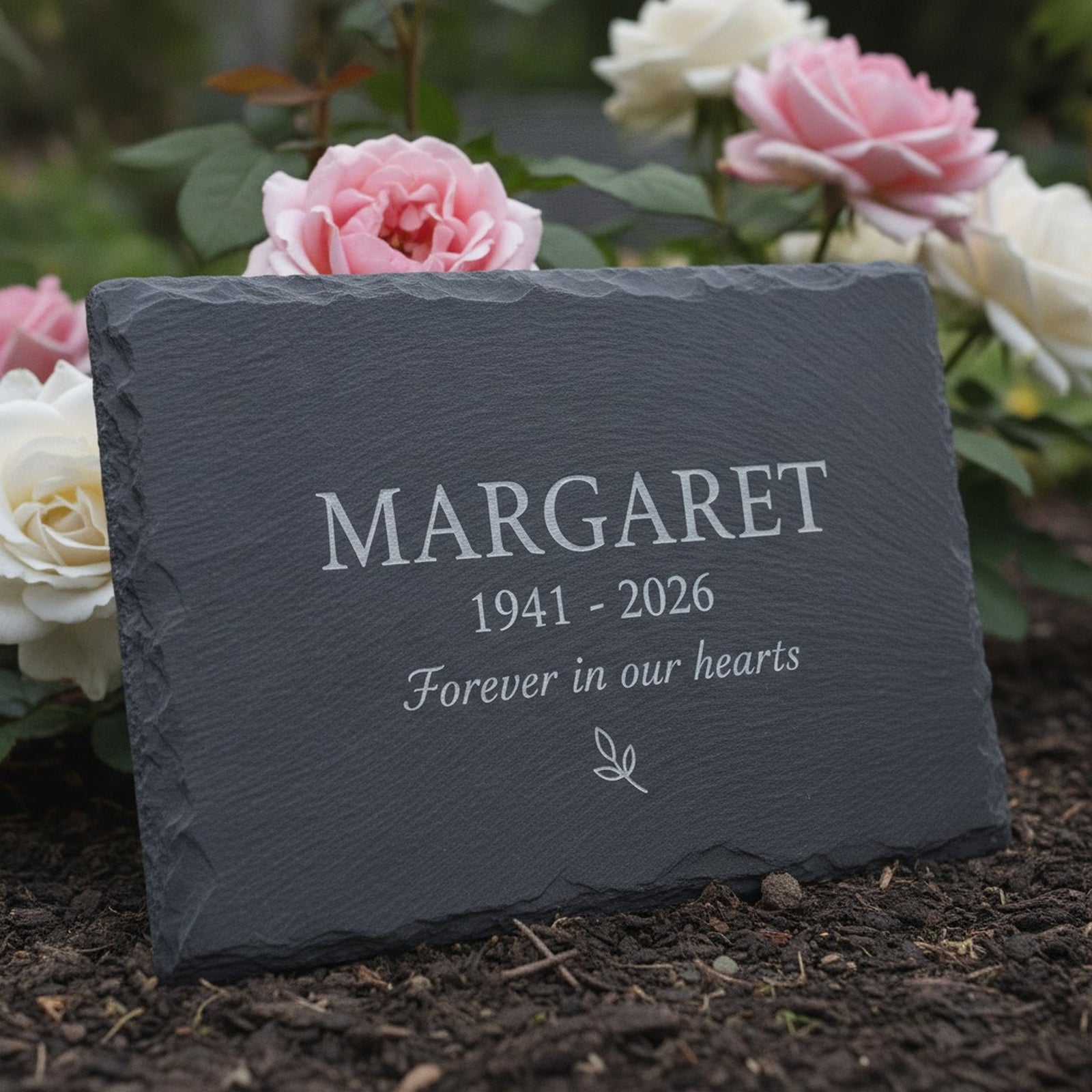 Personalized memorial garden stone in natural slate set in a rose bed, laser engraved with the sample name Margaret, the years 1941 to 2026 and a small leaf sprig