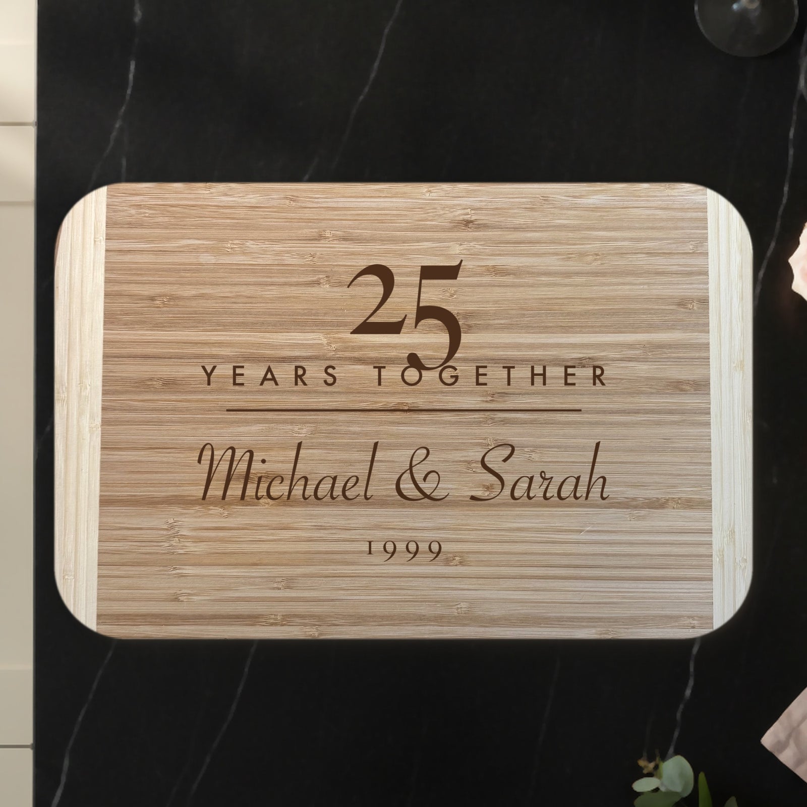 Personalized anniversary cutting board on a black marble countertop with roses and champagne, engraved 25 YEARS TOGETHER above Michael and Sarah on two tone bamboo.
