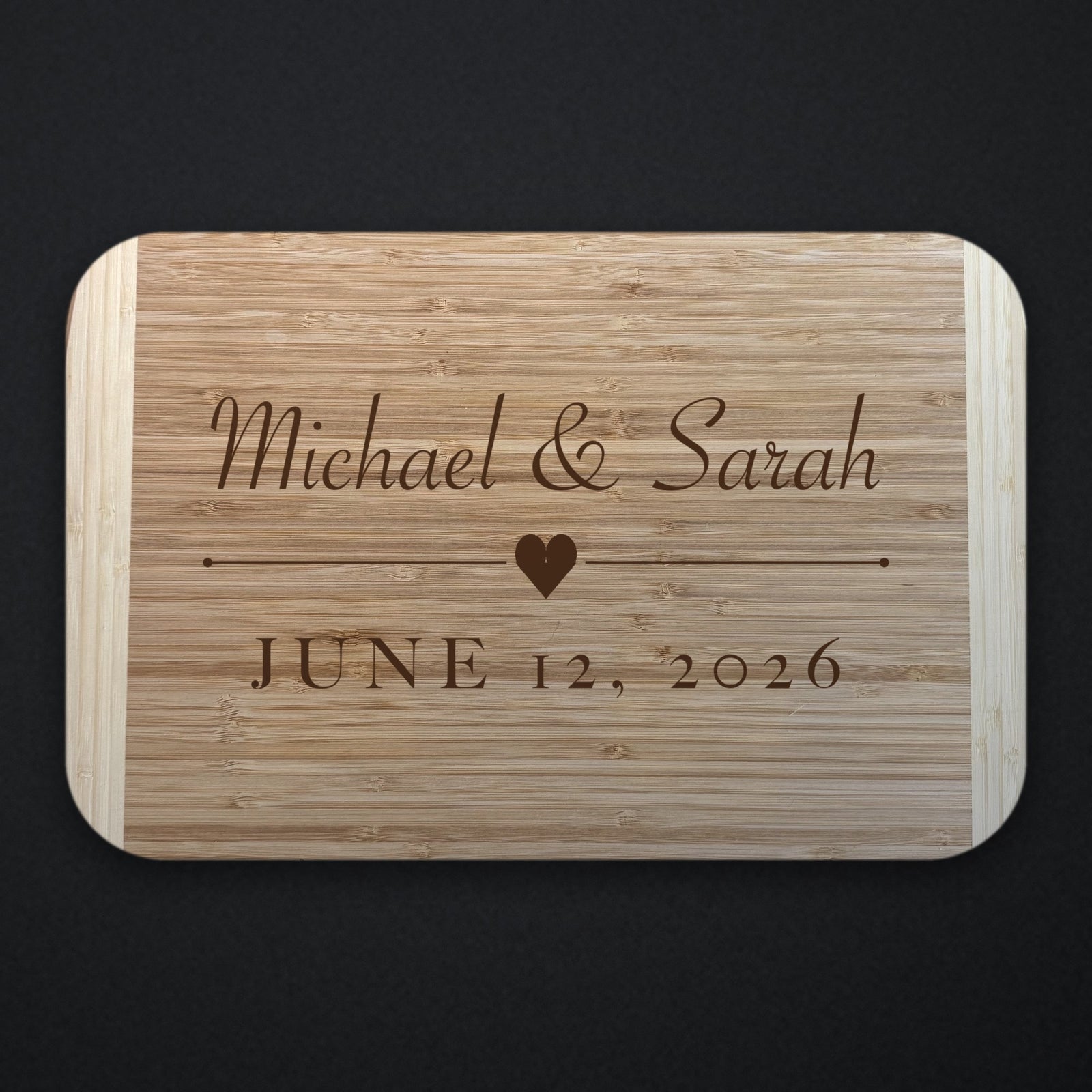 Two tone bamboo couples cutting board engraved with Michael and Sarah in script above a heart and rule, with JUNE 12 2026 beneath, shown on a dark background.