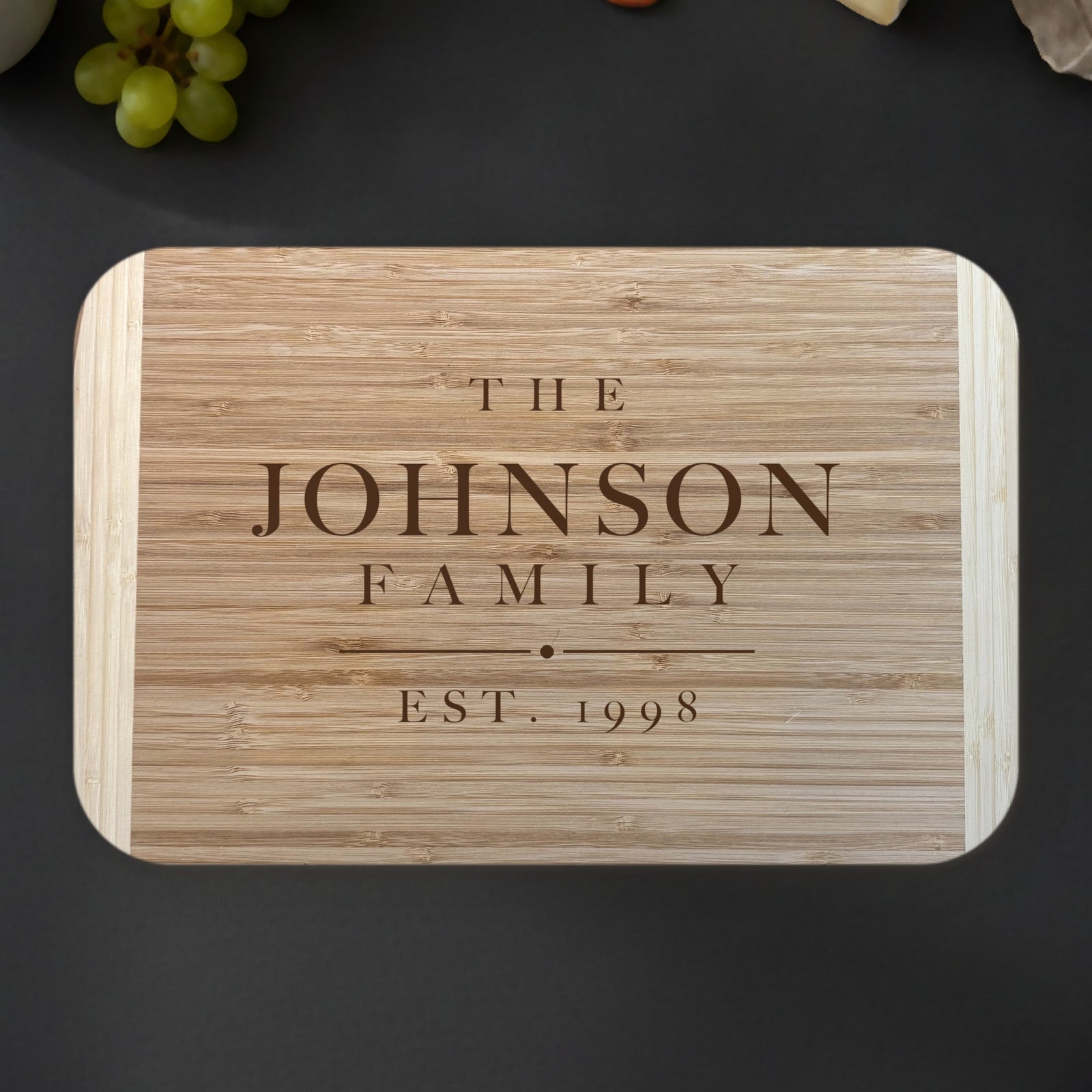 Personalized family name cutting board on a dark slate kitchen counter with grapes and eucalyptus, engraved THE JOHNSON FAMILY EST. 1998 on two tone bamboo.