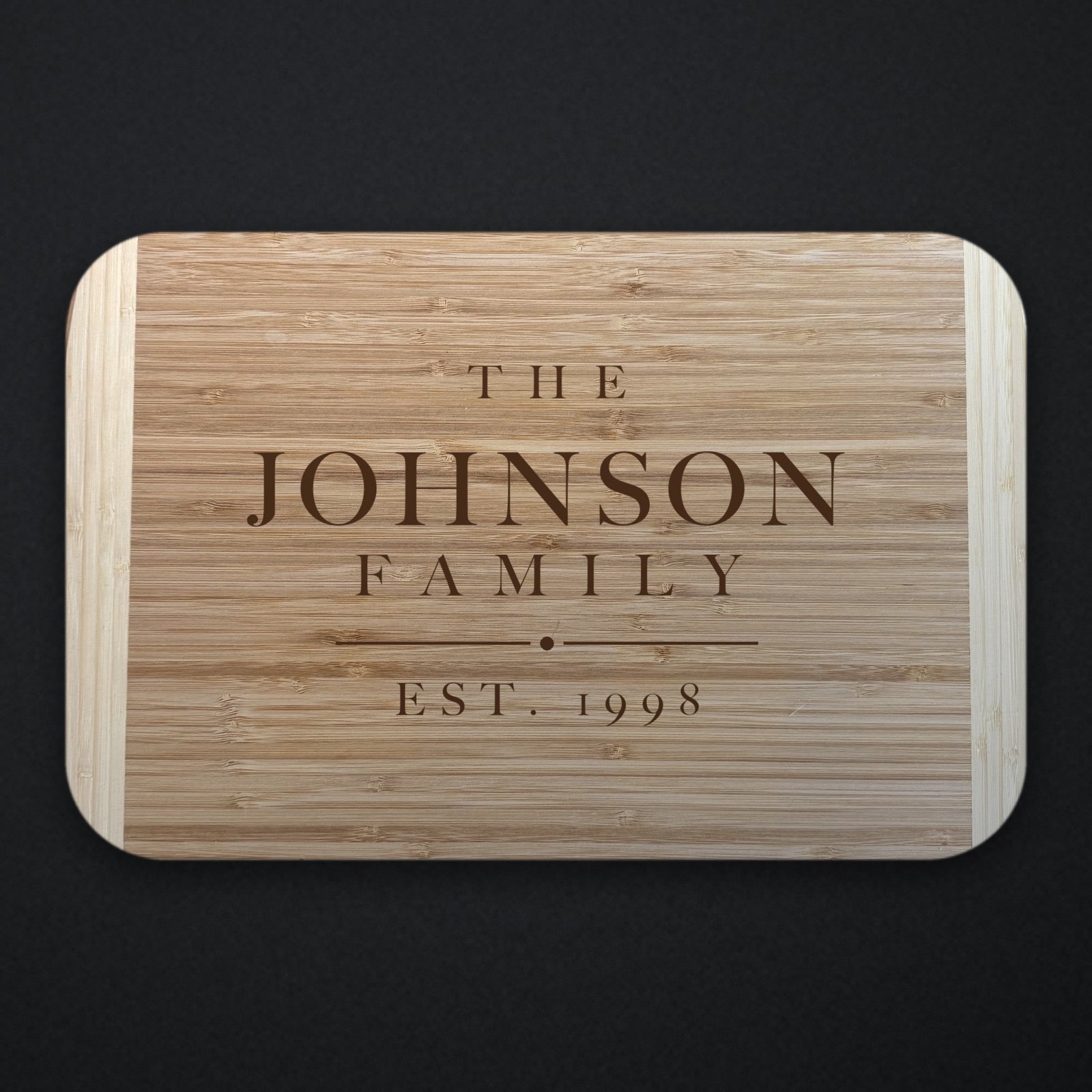 Two tone bamboo cutting board engraved THE JOHNSON FAMILY with EST. 1998 beneath a rule and centre dot, shown on a dark background.