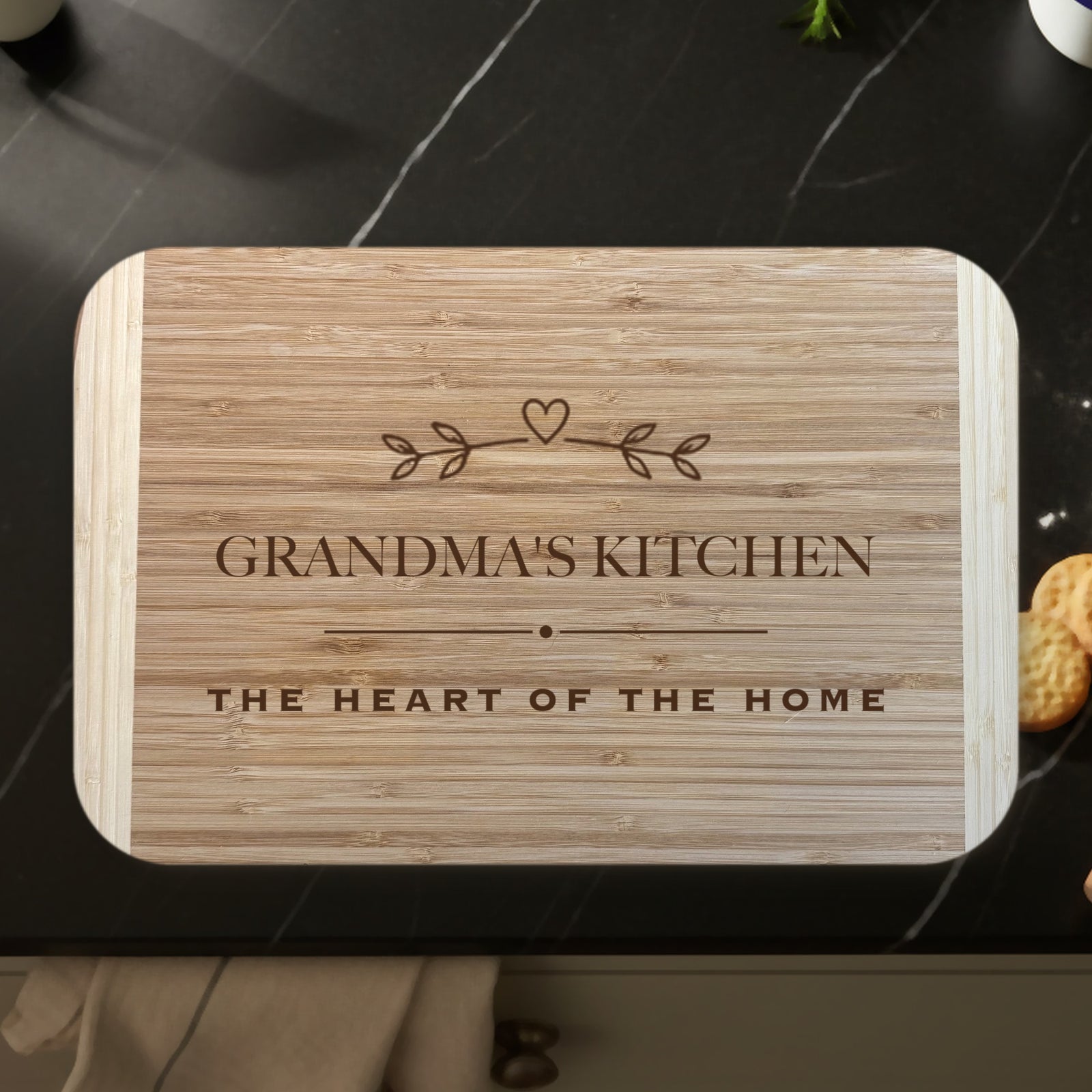 Personalized Grandmas Kitchen cutting board on a black marble countertop, engraved GRANDMA'S KITCHEN and THE HEART OF THE HOME on two tone bamboo.