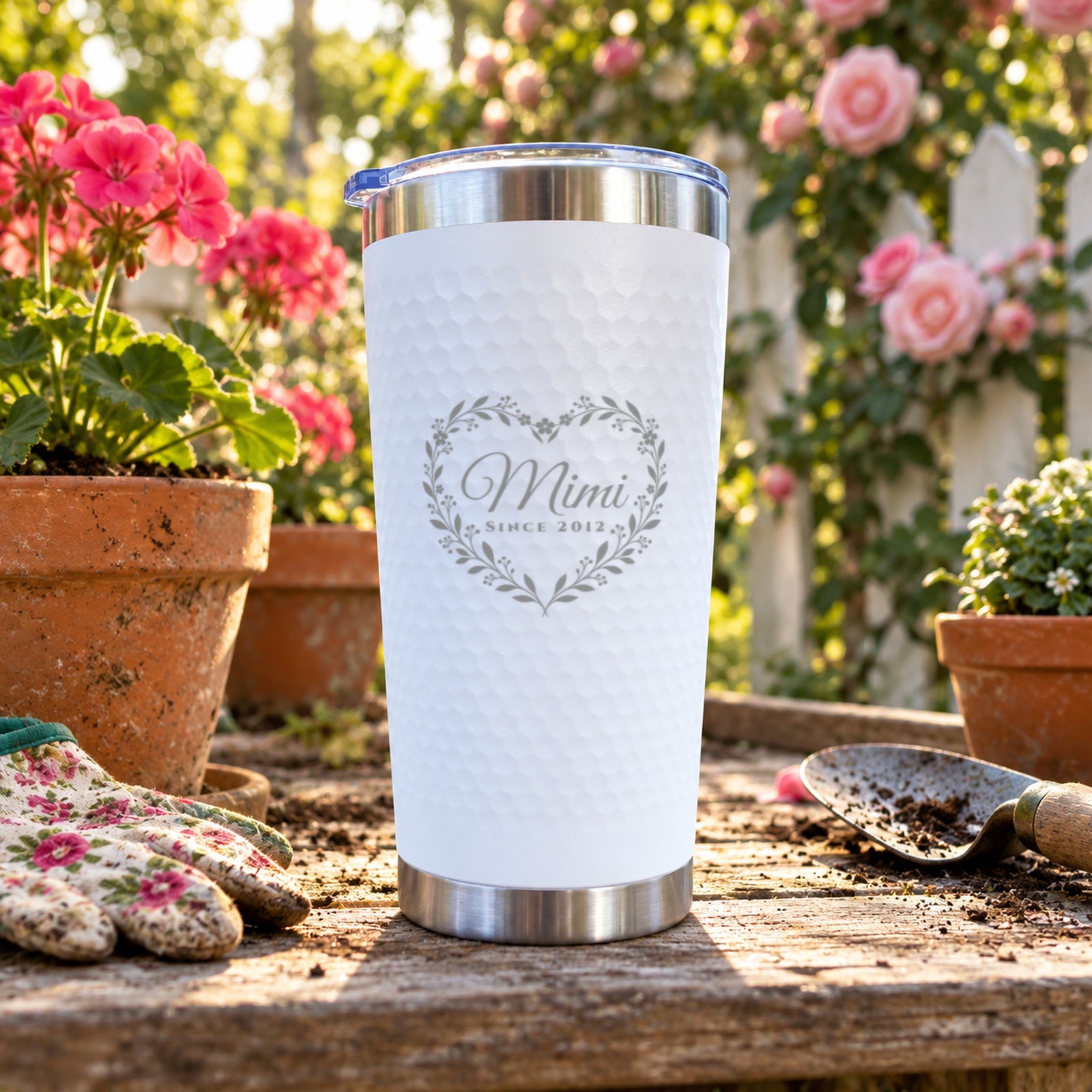 White dimpled tumbler engraved with a heart wreath around Mimi and Since 2012, standing on a garden potting bench beside geraniums