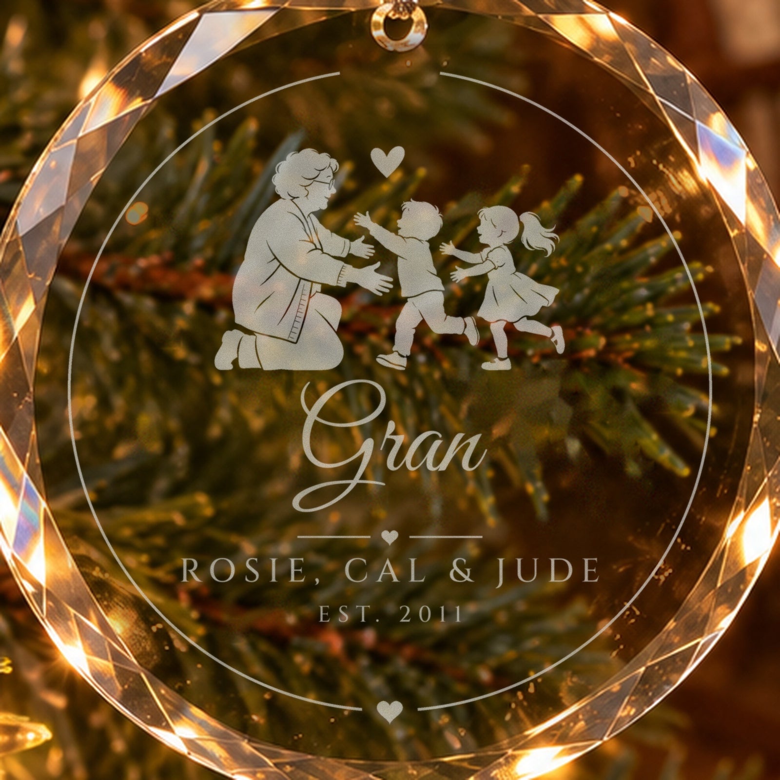 Close-up of the frosted engraving on the grandma ornament: a grandmother opening her arms to two running grandkids, Gran in script, ROSIE, CAL & JUDE, EST. 2011