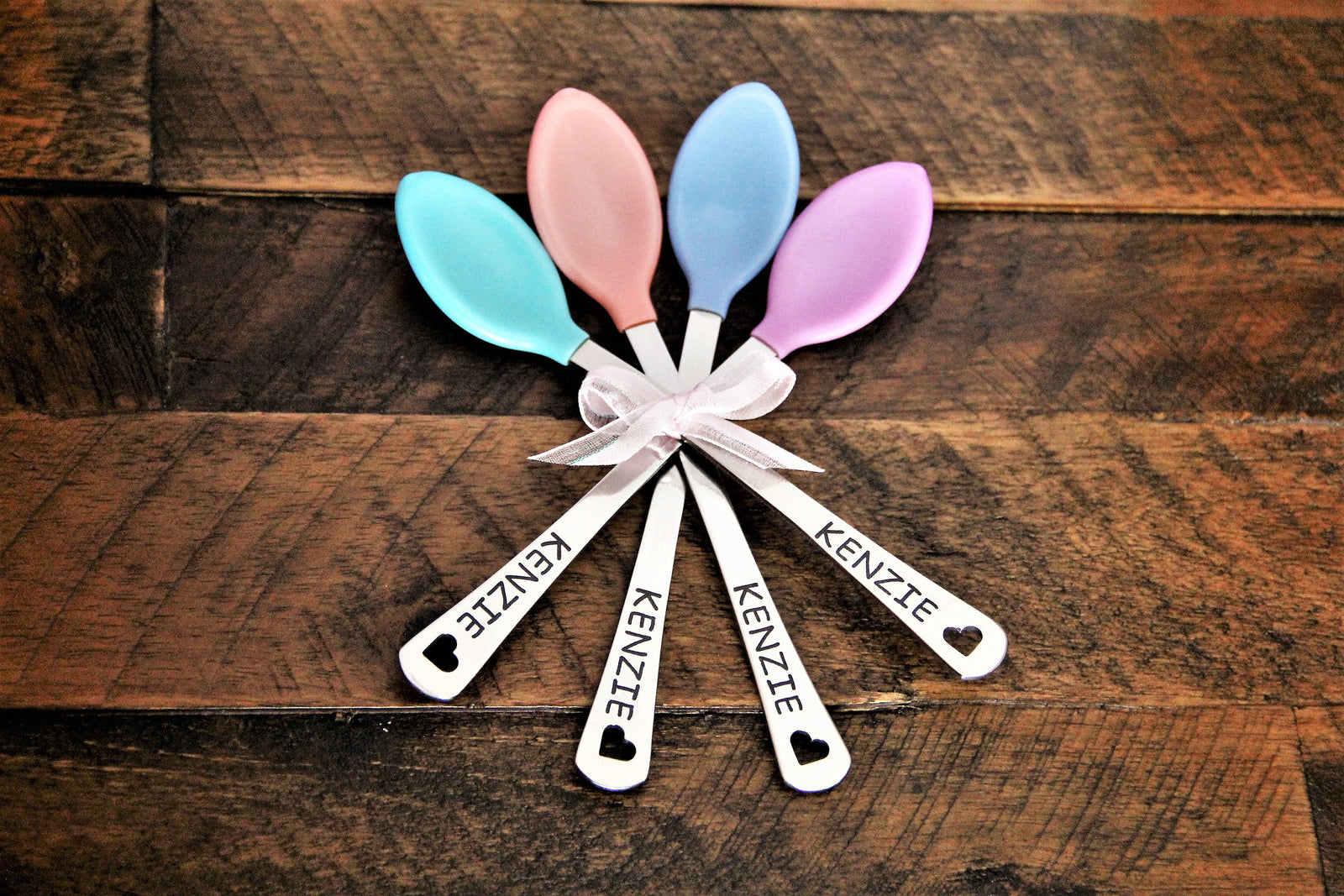 Four personalized baby spoons in pastel pink, peach and lavender silicone, hand-engraved with the sample name Kenzie and tied together with a white ribbon ready for gifting.