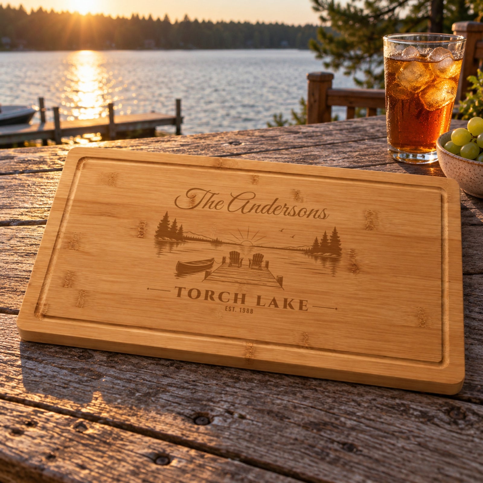 Lake house hostess gift: bamboo cutting board engraved The Andersons, Torch Lake, EST. 1988 with a dock scene, on a deck table by the lake at sunset