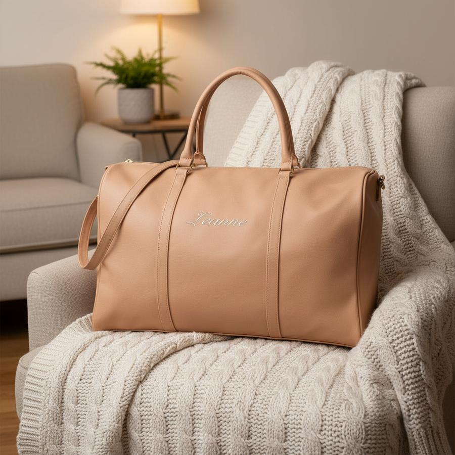Tan vegan leather weekender bag with dual handles displayed on a cream chair, embroidered with sample name "Leanne" on the front panel.