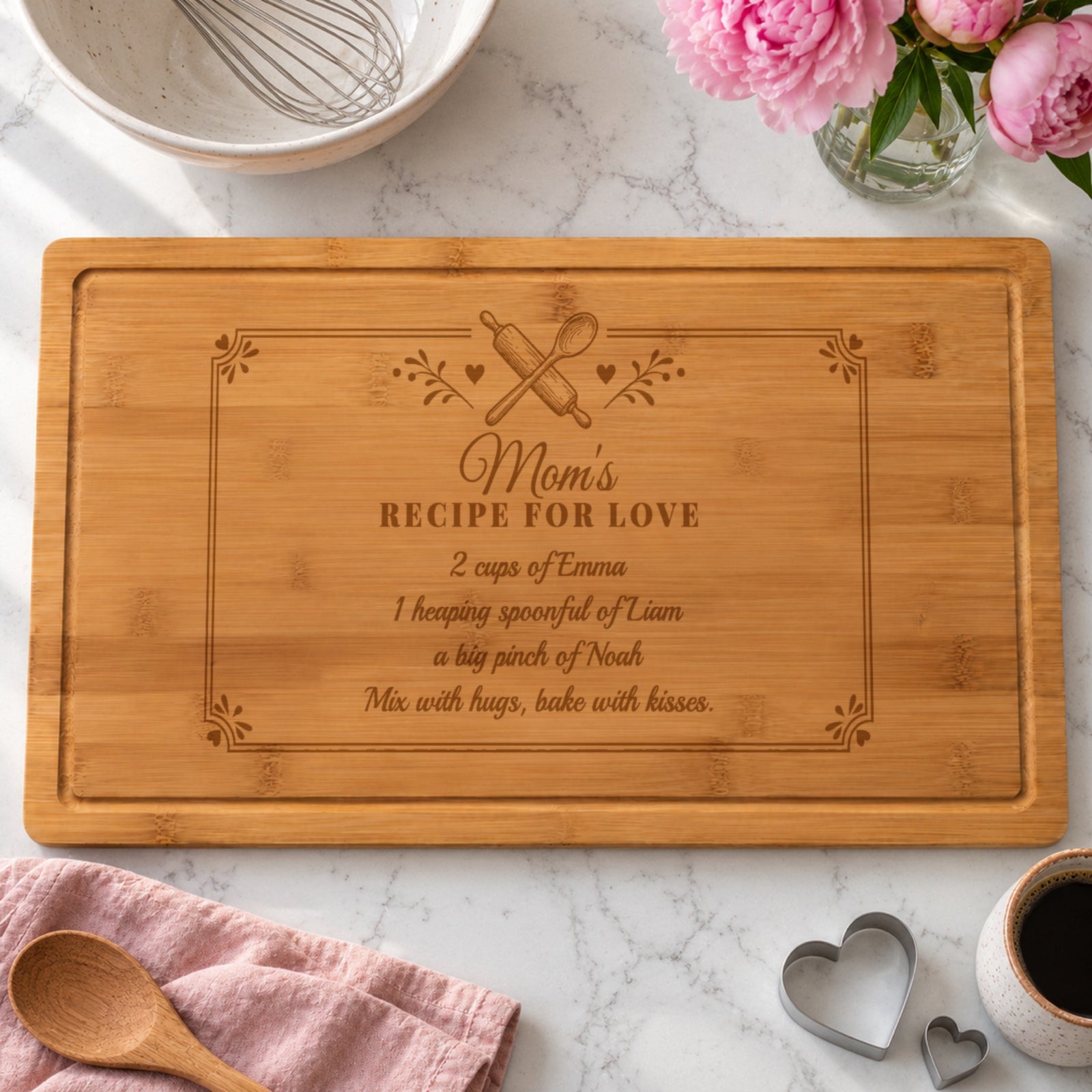 Mom cutting board engraved Mom's Recipe for Love with 2 cups of Emma, 1 heaping spoonful of Liam and a big pinch of Noah, on a white kitchen counter with peonies