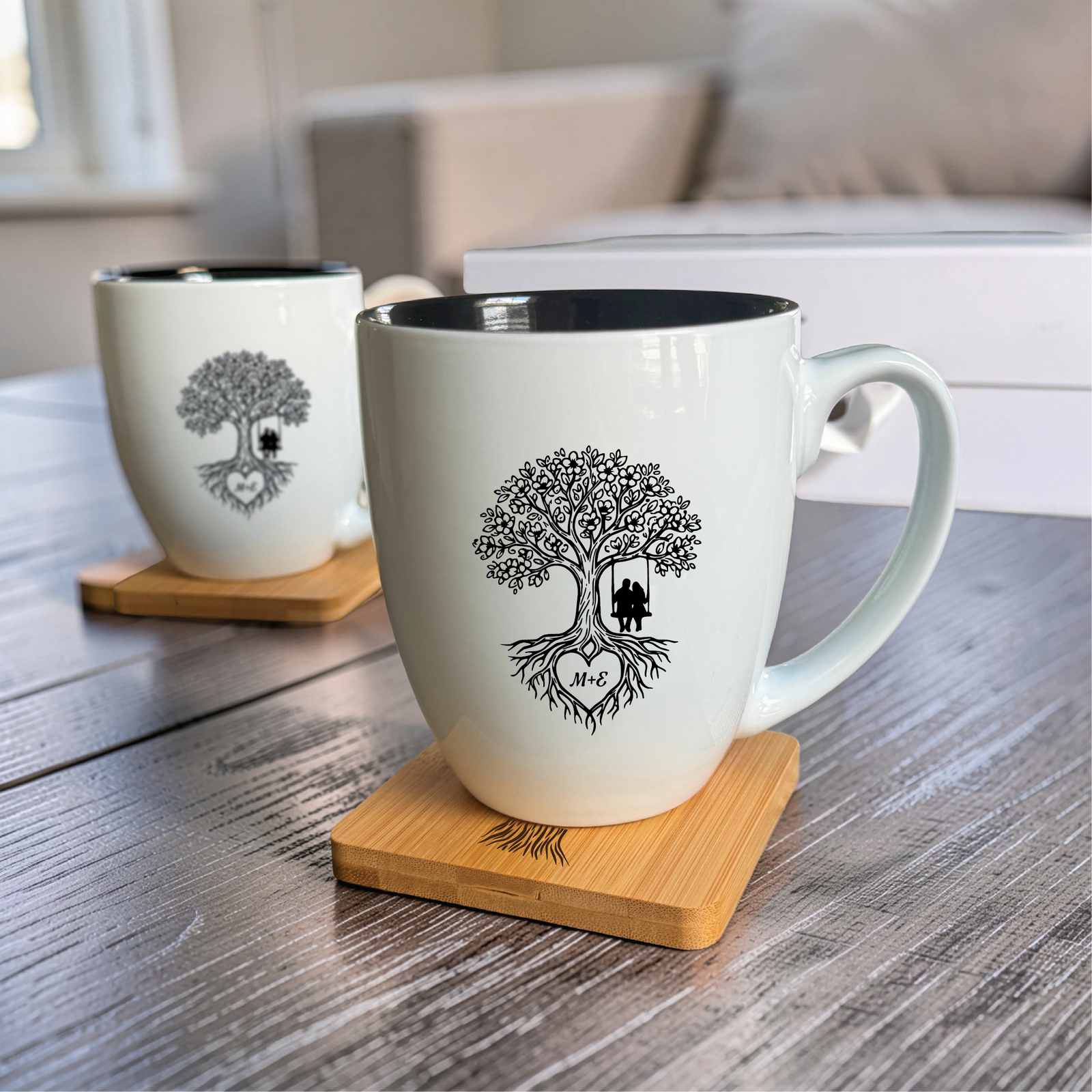 Two 16 ounce white bistro mugs with black interiors engraved with a tree of life and the couple two initials, each resting on a bamboo coaster.