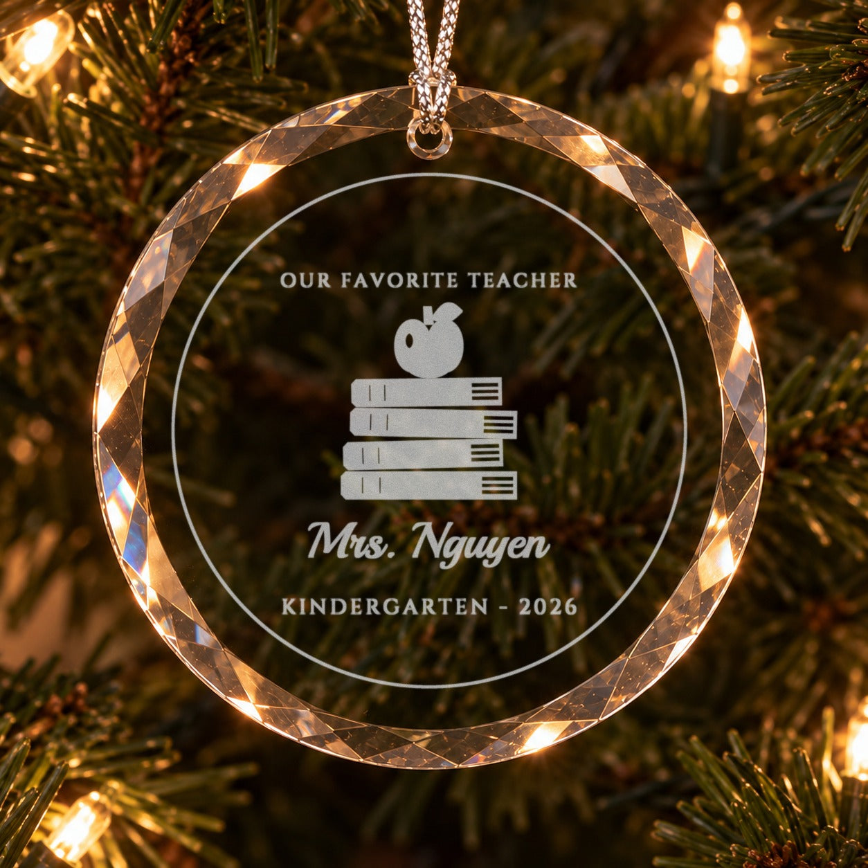 Close-up of the frosted engraving on the teacher ornament: OUR FAVORITE TEACHER, a stack of books with an apple, Mrs. Nguyen in script and KINDERGARTEN 2026