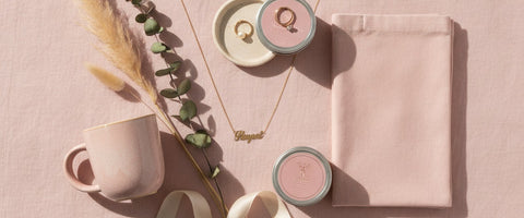 A blush flat-lay of just because gifts under $30 for her including a gold name necklace, ceramic ring dish, candle, mug, and canvas tote.