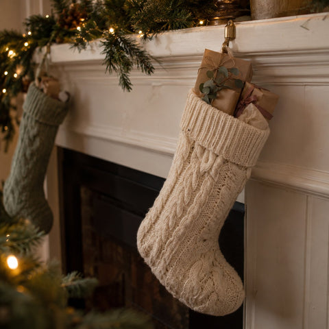 Cream cable knit Christmas stocking hanging from a white mantel, with small kraft paper wrapped stocking stuffers for her tied in twine and a dusty rose ribbon