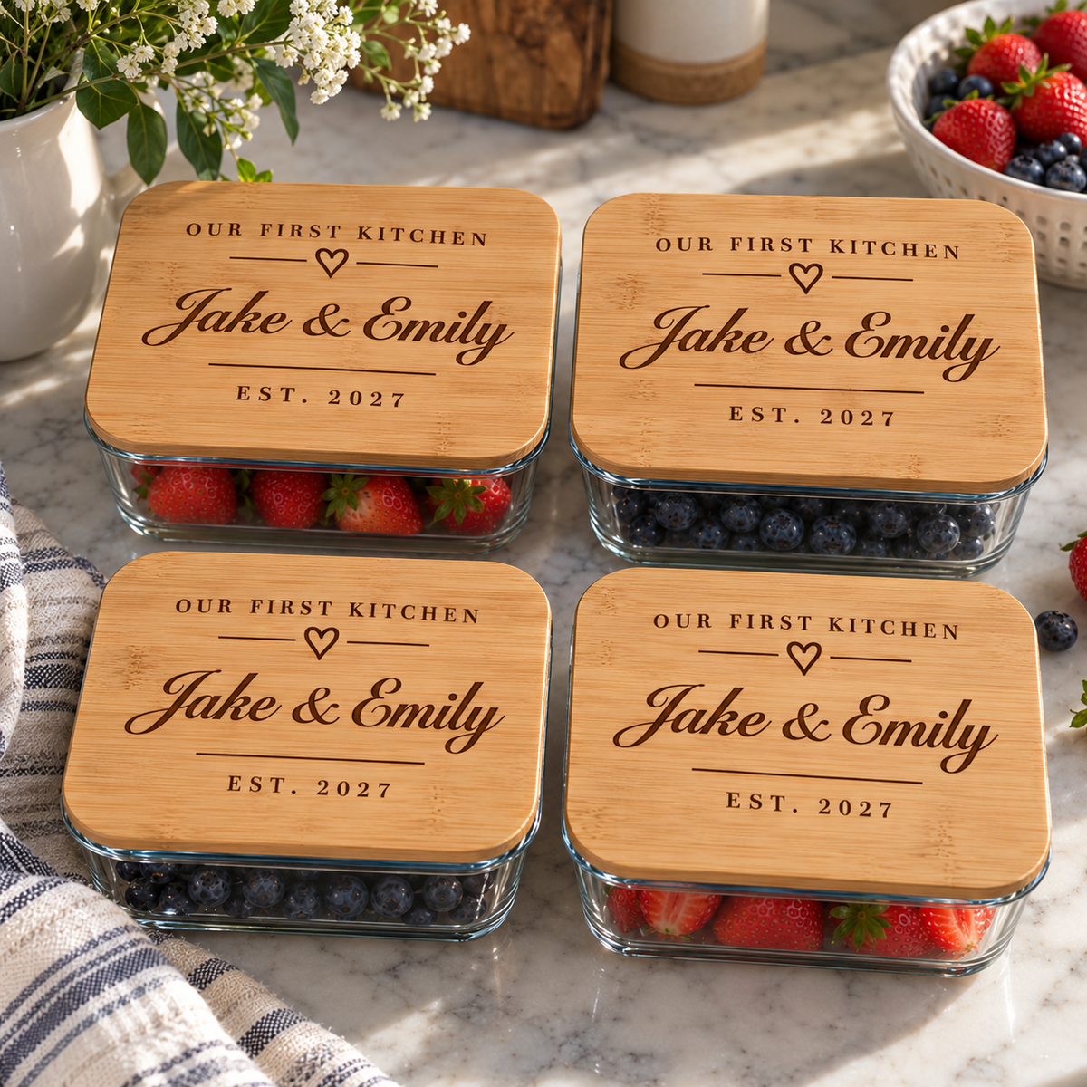 Four glass food storage containers with bamboo lids engraved Our First Kitchen, Jake &amp; Emily, EST. 2027, filled with strawberries and blueberries on marble