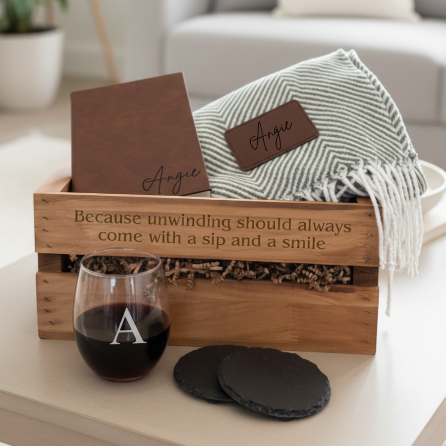 Wooden cedar crate filled with personalized items including a grey striped throw blanket with sample name "Angie" on brown patch, stemless wine glass engraved with sample initial "A", black coasters, and crate engraved with "Because unwinding should always come with a sip and a smile