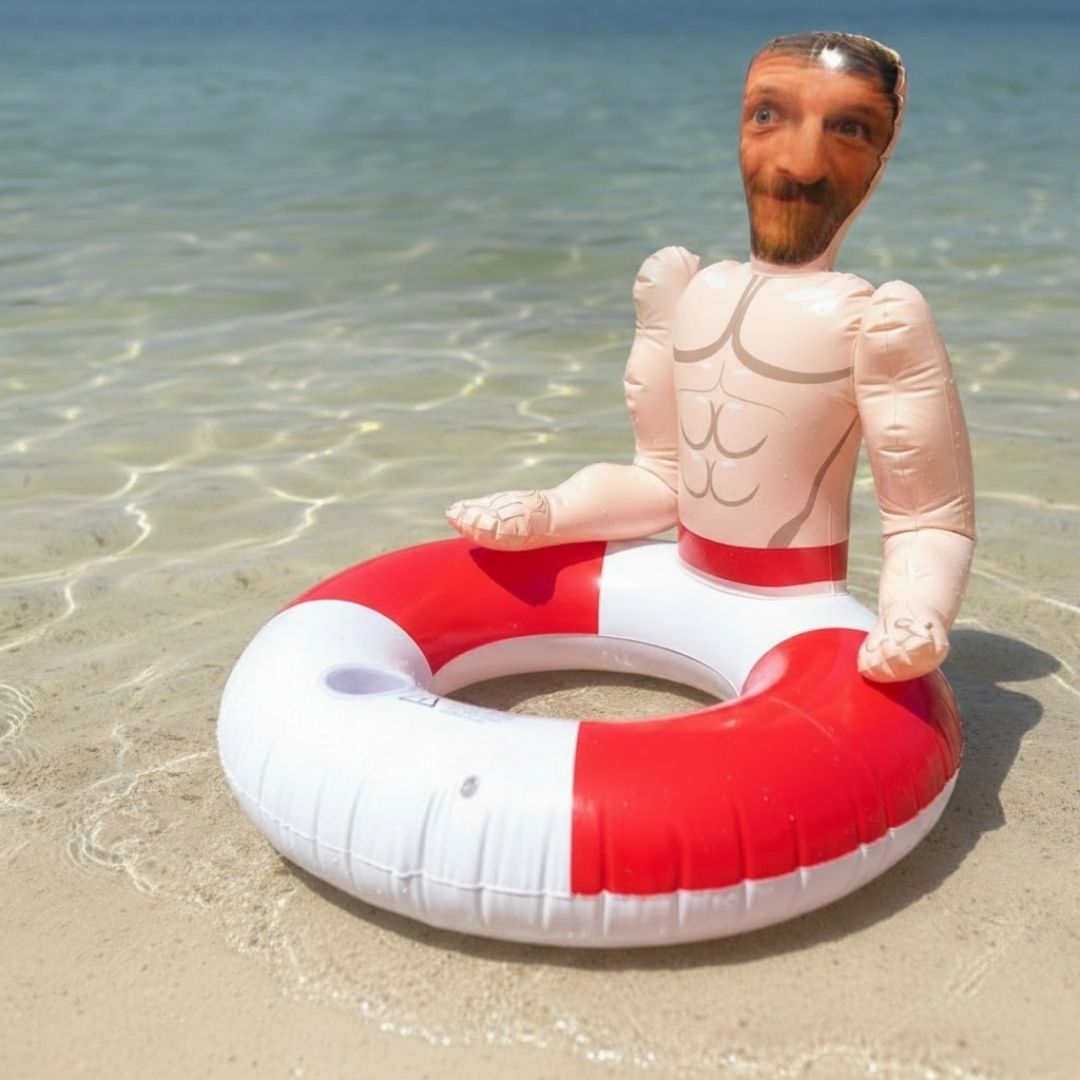 Inflatable pool float with red and white color blocking, featuring a large circular opening and a novelty head and torso attachment with muscular chest detail, positioned in shallow ocean water.
