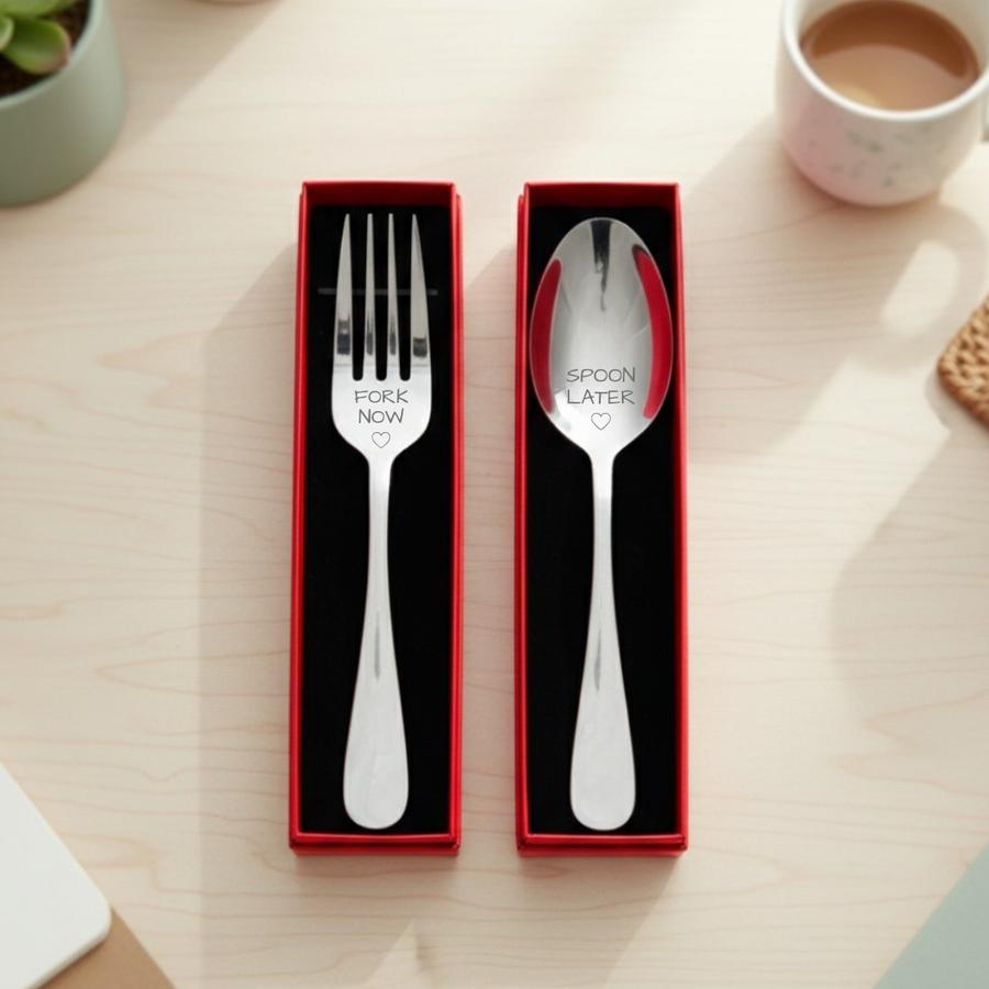 Stainless steel fork engraved "FORK NOW" and spoon engraved "SPOON LATER" with small hearts, displayed in red gift boxes with black interiors on a wooden surface.