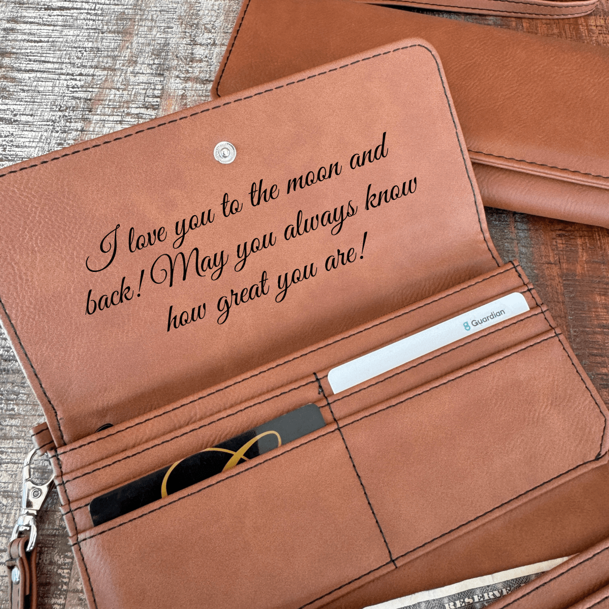 Brown personalized wristlet open flat showing the engraved inside flap message, card slots and interior pockets on a rustic wood table.