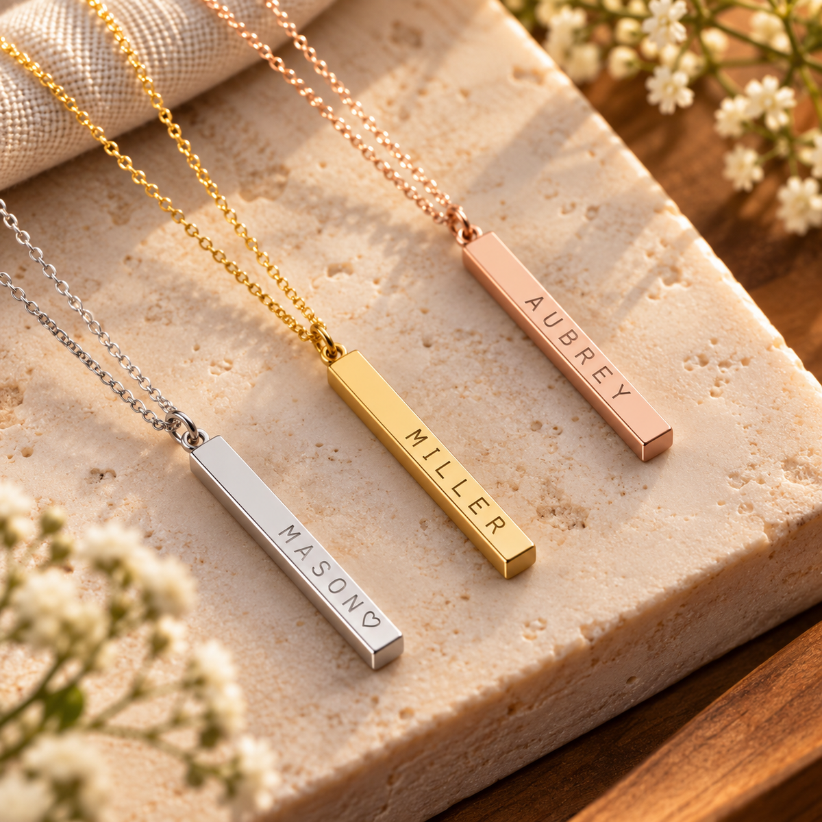 Three personalized bar name necklaces in silver, gold, and rose gold are displayed on a textured beige stone slab with small white flowers. The necklaces have engraved names: MASON, MILLER, and AUBREY.