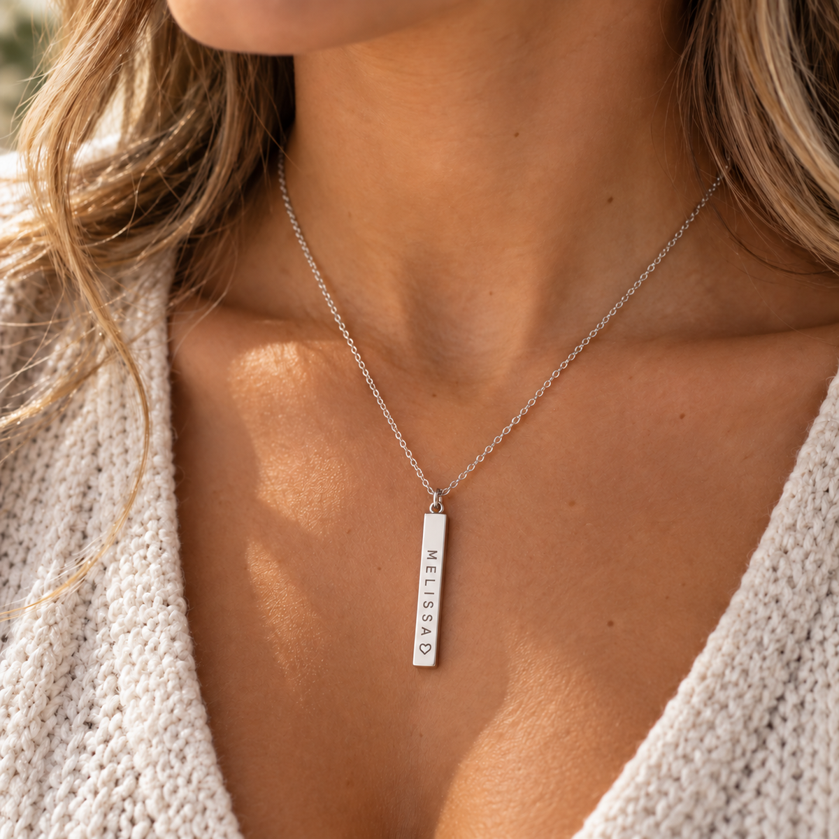 A close-up of a woman wearing a delicate silver chain necklace with a vertical bar pendant engraved with the name 'MELISSA'. She is wearing a cream-colored knit sweater, and the background is softly blurred.
