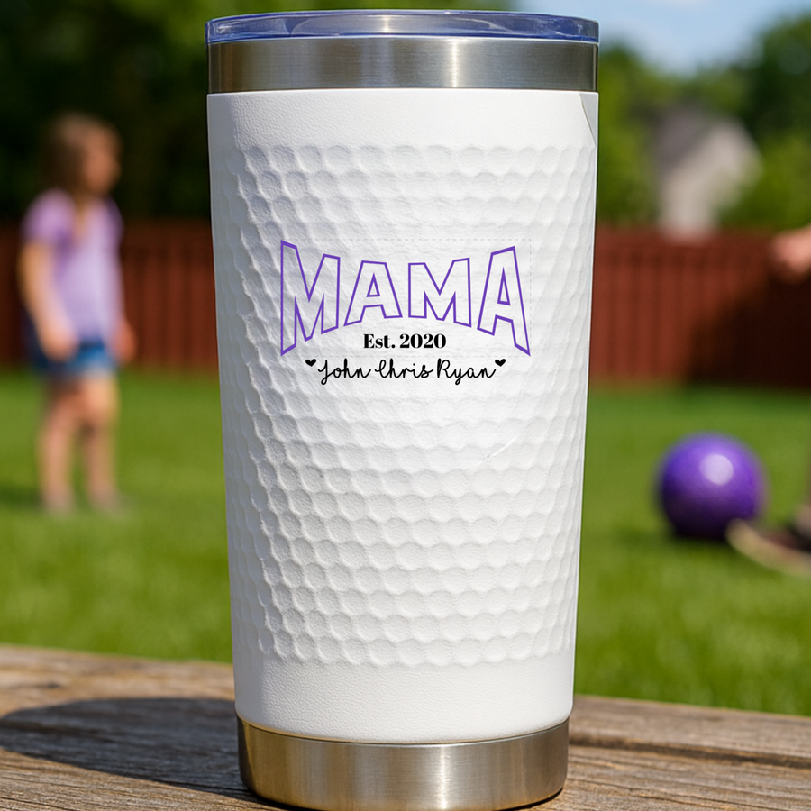White textured tumbler with stainless steel band featuring purple "MAMA" text, sample engraving "Est. 2020" and children's names "John Chris Ryan" with small hearts, displayed outdoors on wooden surface.