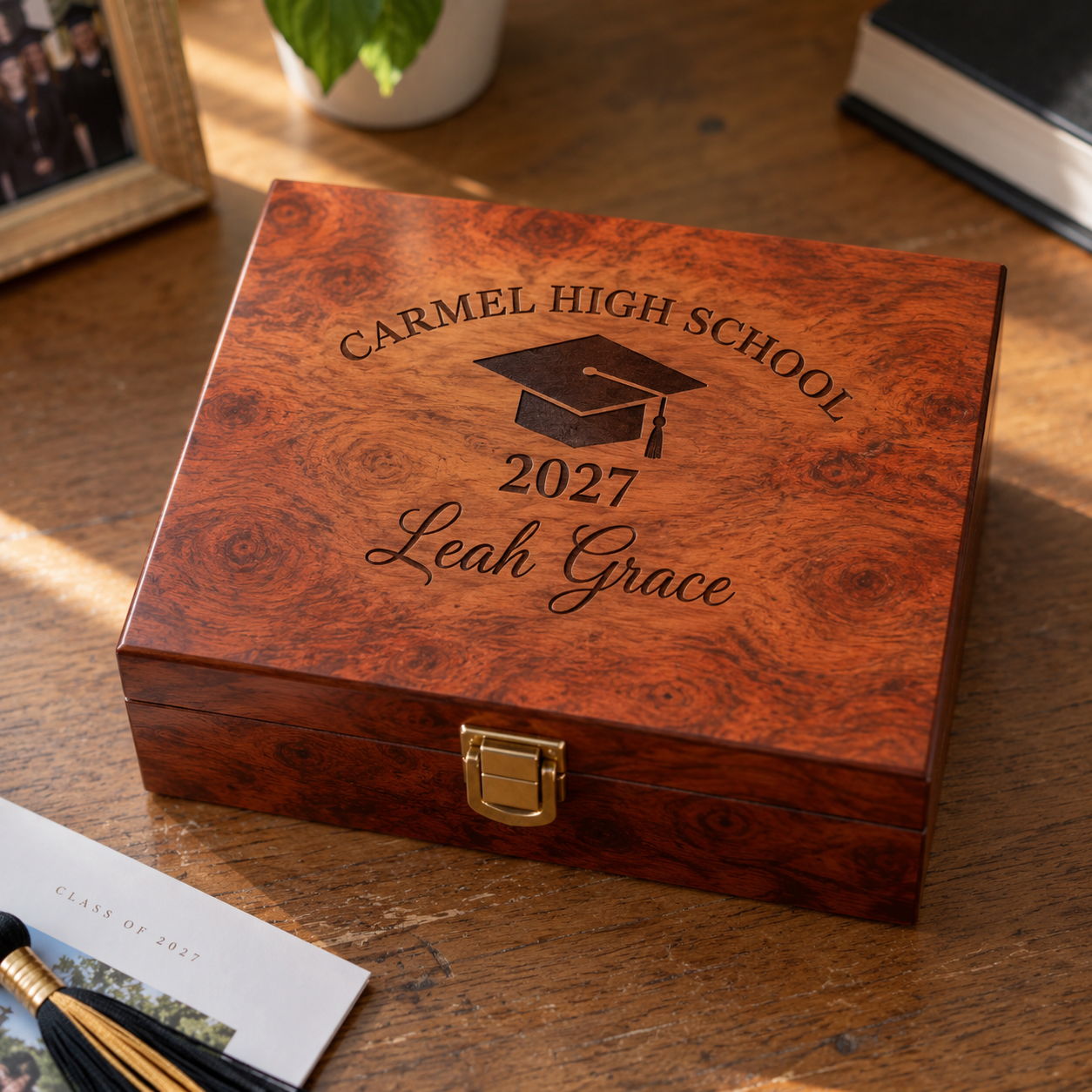 Burl wood graduation keepsake box engraved with Carmel High School, a graduation cap, 2027 and Leah Grace, closed on a sunlit desk beside a class of 2027 card