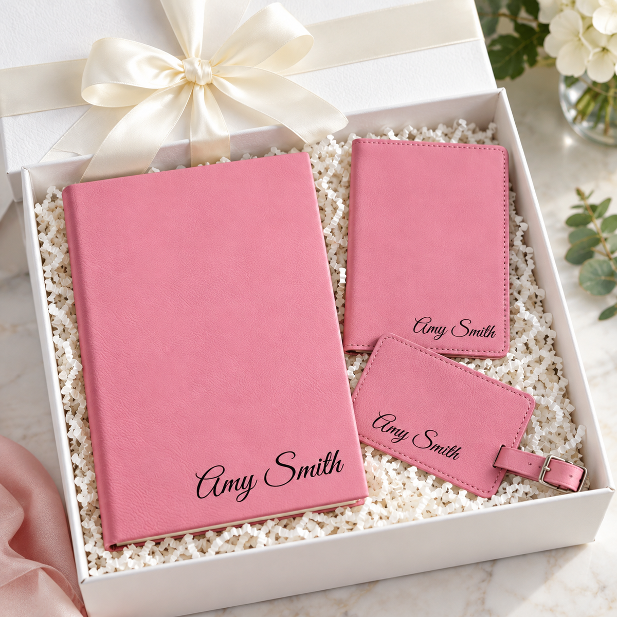Pink leatherette journal, passport holder, and luggage tag personalized with the name Amy Smith, laid in a white gift box with cream ribbon and crinkle paper.