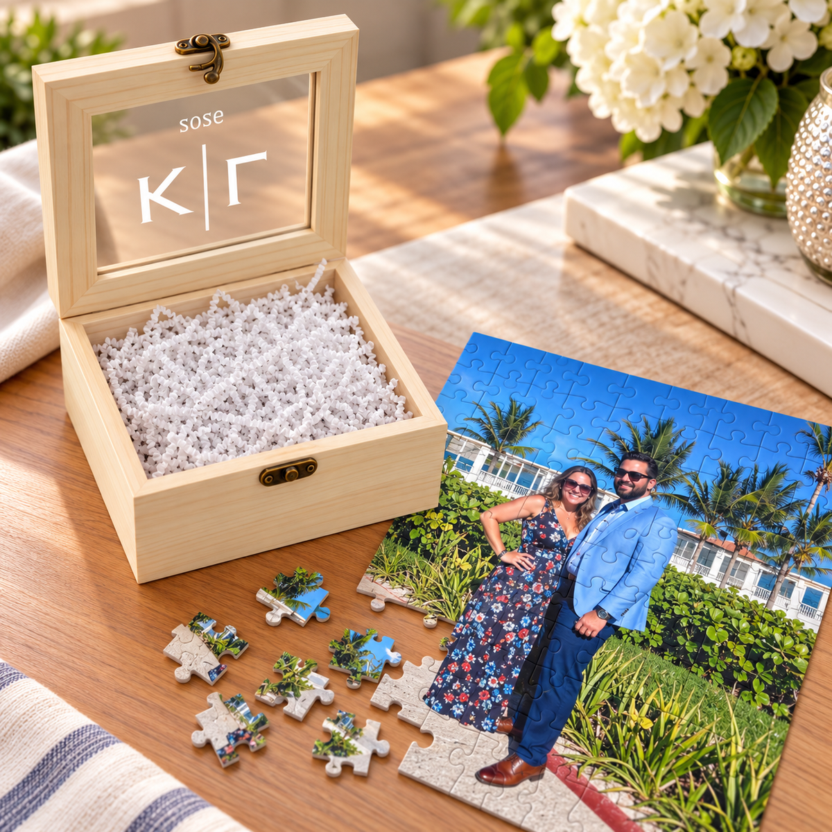 Open wood gift box with a personalized glass window lid, next to an assembled photo puzzle of a couple on a tropical beach with palm trees and loose puzzle pieces