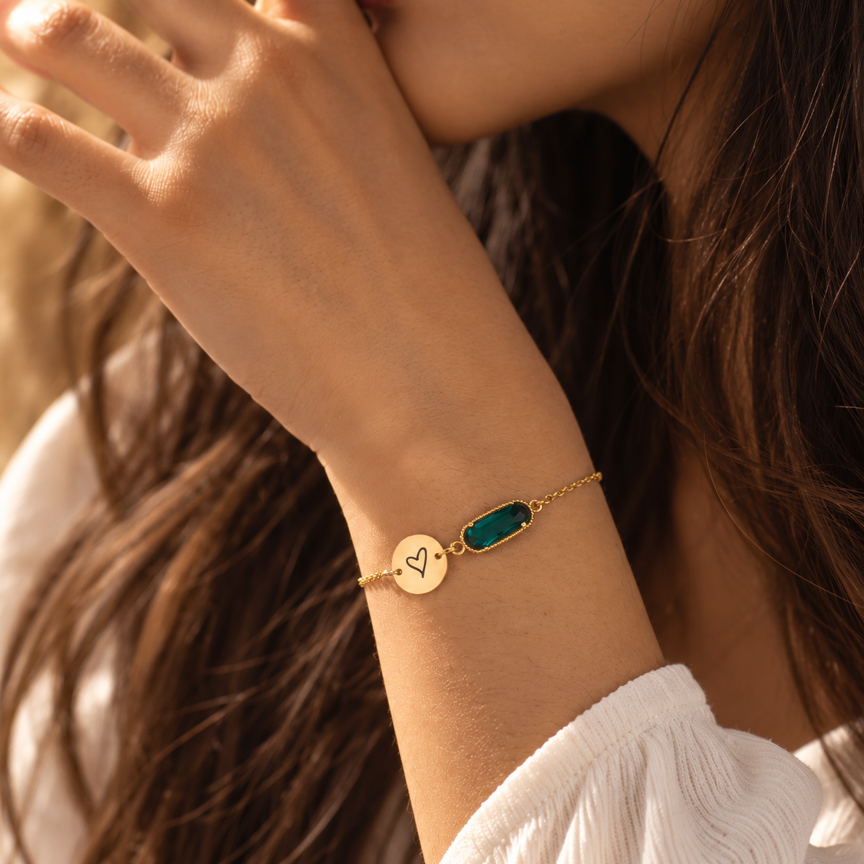 Gold bracelet with teal birthstone and gold charm engraved with a heart, worn on model's wrist against light background